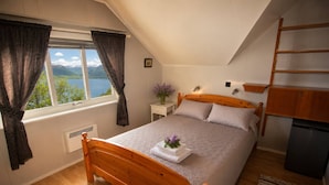Un dormitorio con cama de madera, una ventana con cortinas, una mesita de noche, un radiador, una pequeña mesa con un jarrón y vistas a montañas y un lago.
