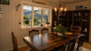 Un comedor con una mesa de madera, sillas, una lámpara de araña, una estantería y una ventana con vistas a montañas.