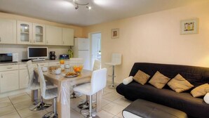 Una cocina moderna con armarios blancos, un comedor con mesa y sillas, un sofá negro con cojines y una televisión.