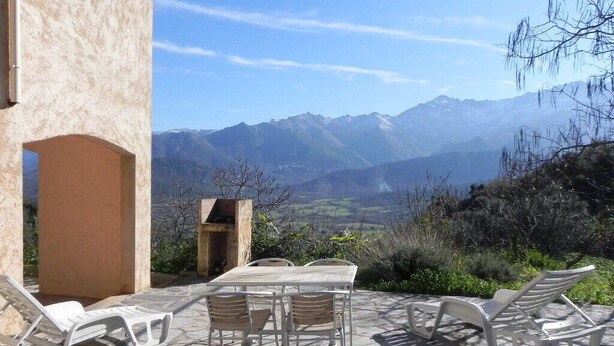 Un patio con suelo de piedra, sillas reclinables blancas y una mesa. Al fondo, montañas y cielo despejado.