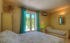 Ein Schlafzimmer mit Bett, Nachttisch, Kommode, einem Fenster mit Vorhängen, einer Klimaanlage und einem Spiegel an der Wand.