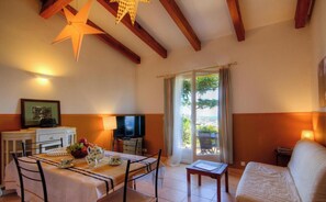 Ein Zimmer mit Esstisch, Stühlen, Fernseher, einer Couch und einem Balkon mit Vorhängen.