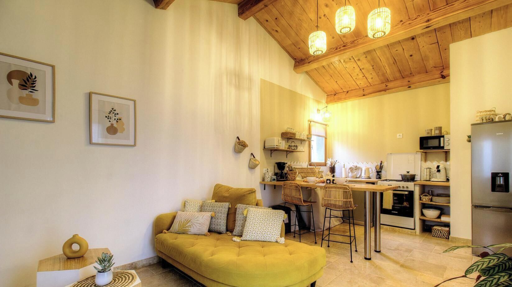 A cozy living room with a yellow sofa, a kitchen with a dining area, and wooden ceiling beams.