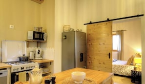 Eine kompakte Küche mit Herd, Mikrowelle und Kühlschrank. Eine Holztür führt zu einem Schlafzimmer mit Bett und Nachttisch.