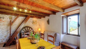 Un'area pranzo rustica con soffitto in legno, un tavolo apparecchiato con tovaglia gialla, un cesto di vimini con fiori e vista sulla campagna attraverso una finestra.