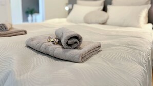Cama bem arrumada com toalha dobrada e um objeto pequeno em cima, cabeceira com almofadas e porta que dá para outro quarto.