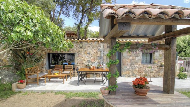 Un edificio de piedra con una pérgola de madera, mesas al aire libre y plantas en macetas.