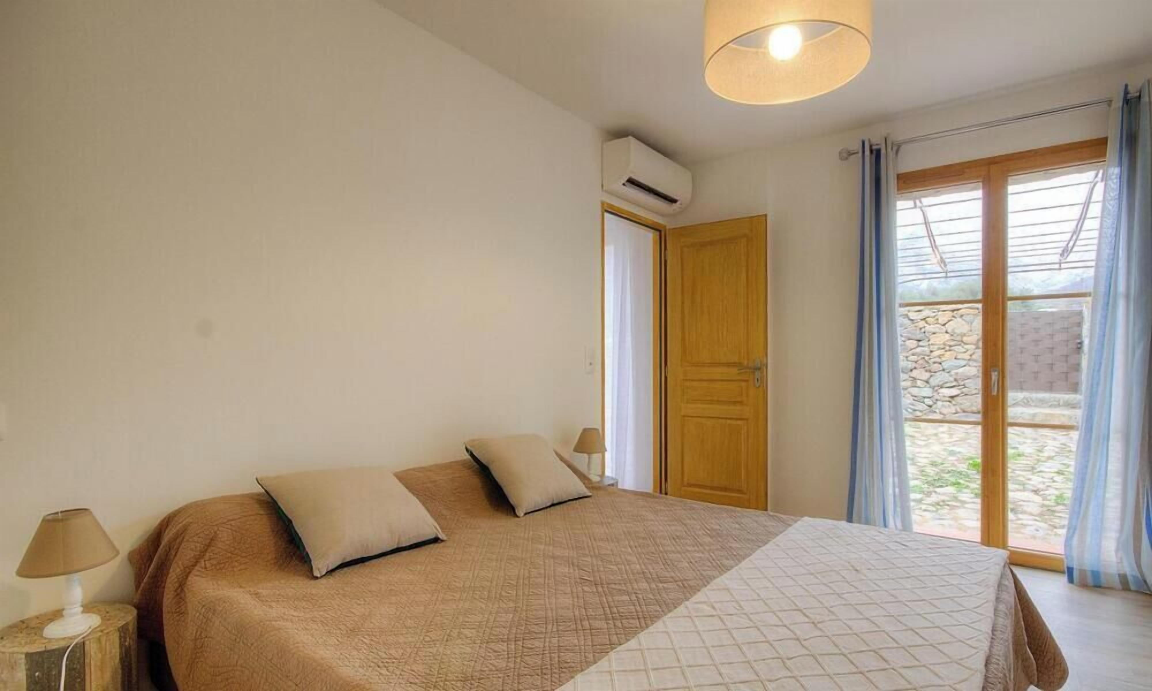 Ein Schlafzimmer mit einem Bett, zwei Kissen, einer Nachttischlampe, einer Klimaanlage, einer Holztür und einem Blick durch ein Fenster mit Vorhängen.