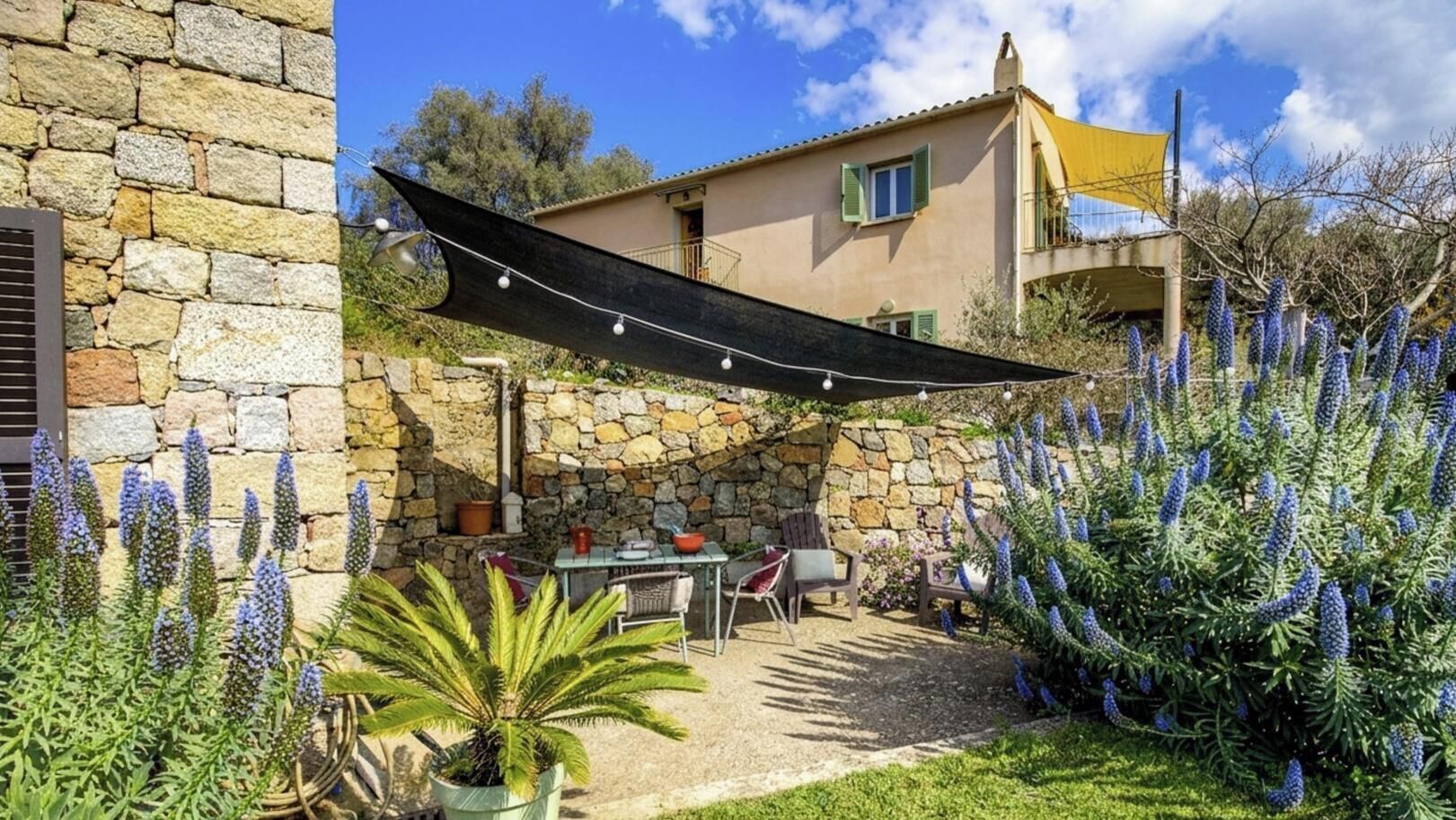A stone house with a patio, outdoor seating, and a hanging shade.
