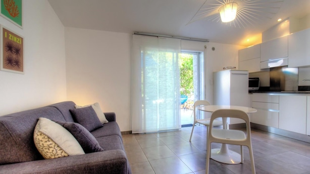 Ein modernes Wohnzimmer mit einer Couch, einem Esstisch mit Stühlen, einer Küchenzeile mit Schränken und einer Glasschiebetür, die zu einem Außenbereich führt.