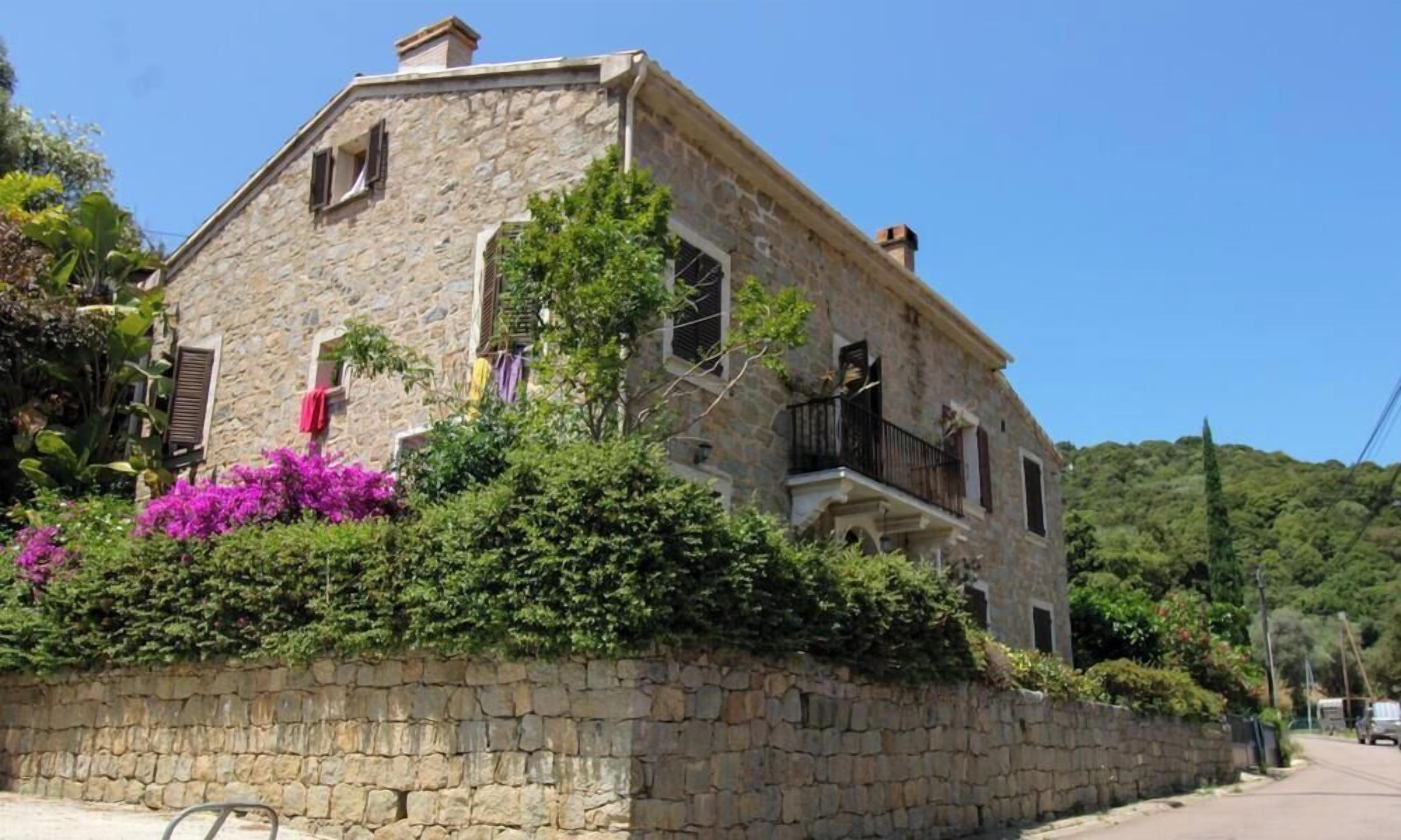 Um edifício de pedra com varanda, cercado por vegetação exuberante e flores.