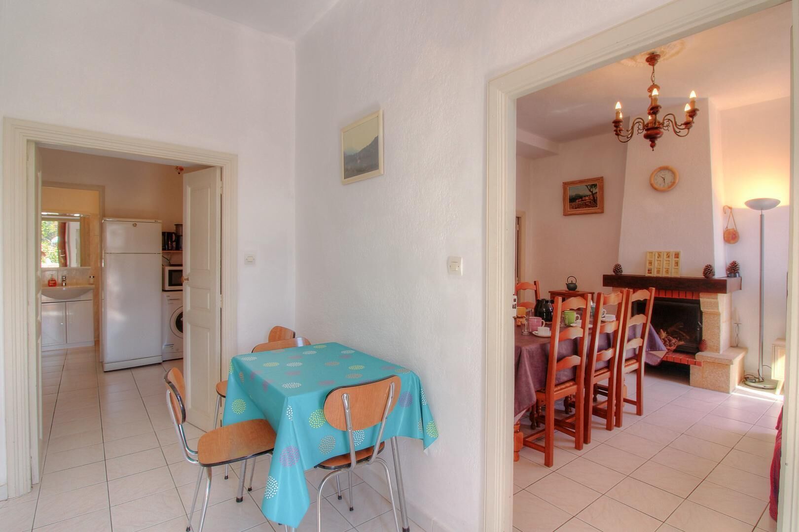 A dining area with a table set for four, a kitchen in the background, and a fireplace with a clock on the wall.