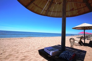 Eine Strandansicht mit einer Holzbank, einem Tisch mit einer Kiste und einem Kissen sowie einem roten Sonnenschirm.