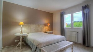Ein Schlafzimmer mit einem Bett, Nachttischlampen, einem kleinen Tisch, einer Bank und einem Fenster mit Vorhängen.