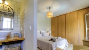 Un baño con lavamanos, espejo y toallero. La habitación tiene un armario de madera y una cama con edredón y almohadas blancas.