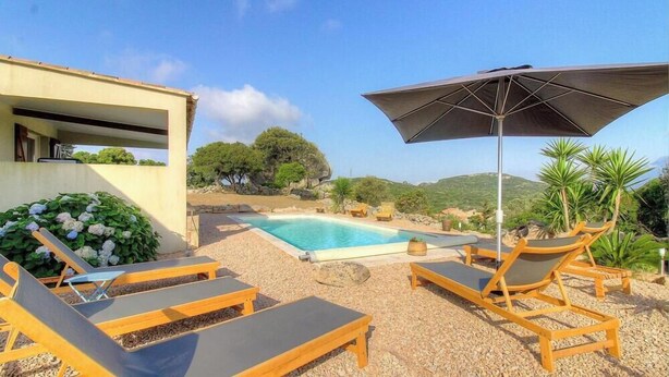 Zona de piscina con sillas de descanso y una sombrilla, rodeada de vegetación y colinas.