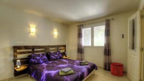 Ein Schlafzimmer mit einem hölzernen Kopfende, zwei Nachttischlampen, einem Bett mit violettem Überwurf, einem Fenster mit Vorhängen und einem roten Mülleimer.