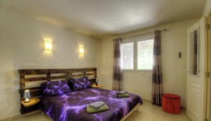 Ein Schlafzimmer mit einem hölzernen Kopfende, zwei Nachttischlampen, einem Bett mit violettem Überwurf, einem Fenster mit Vorhängen und einem roten Mülleimer.