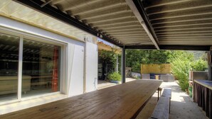 Eine Terrass mit Holztisch und -bänken, eine Glastür zum Küchenbereich und ein überdachter Bereich mit Blick ins Grüne.