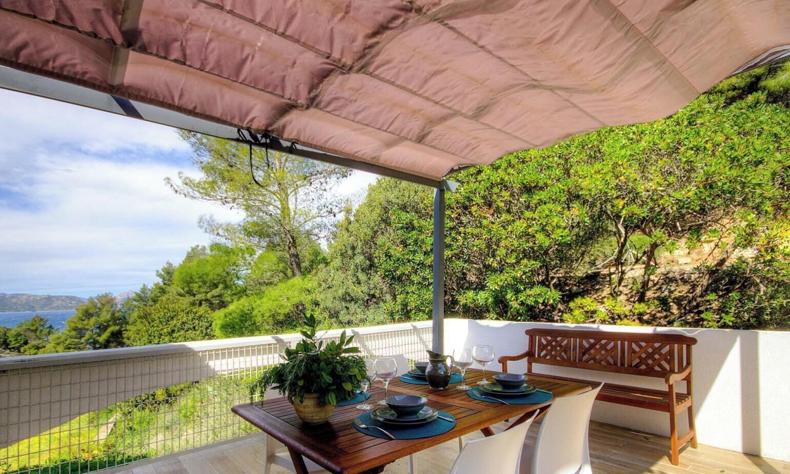 A covered outdoor dining area with a table set for two, a potted plant, and a view of the sea and greenery.
