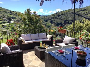 Eine Terrasse mit Korbmöbeln, einem Esstisch und einem blühenden Ampelfleur.