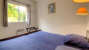 Un dormitorio con cama, ventana con cortinas, radiador, un cuadro en la pared y una lámpara.