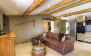 Ein Wohnzimmer mit braunem Ledersofa, Holzbalken an der Decke, einem Holztisch und einer Küchenzeile mit modernen Geräten.