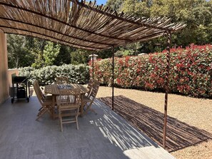 Eine Terrasse im Freien mit Tisch und Stühlen, umgeben von einem Garten mit roten Blättern und einer darüberliegenden Pergola.