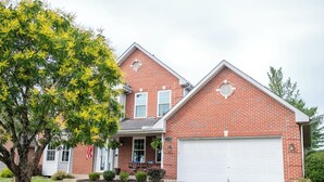 A two-story brick house with a white garage door, a front porch with a bench, and a tree in the yard. - Large Family Home with Backyard Oasis & Fire Pit (Florence)