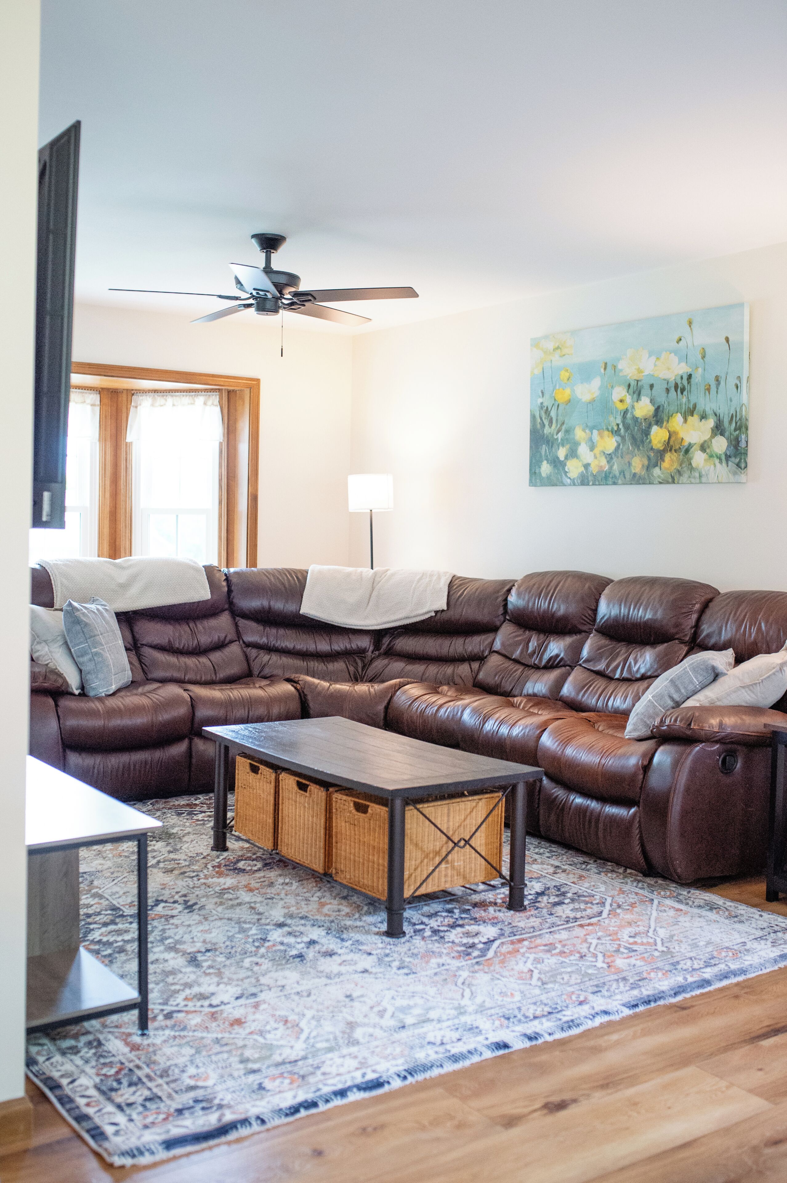 A living room with a brown leather sectional sofa, a wooden coffee table, a patterned rug, a ceiling fan, and a painting of flowers on the wall.