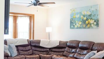 A living room with a brown leather sectional sofa, a wooden coffee table, a patterned rug, a ceiling fan, and a painting of flowers on the wall.