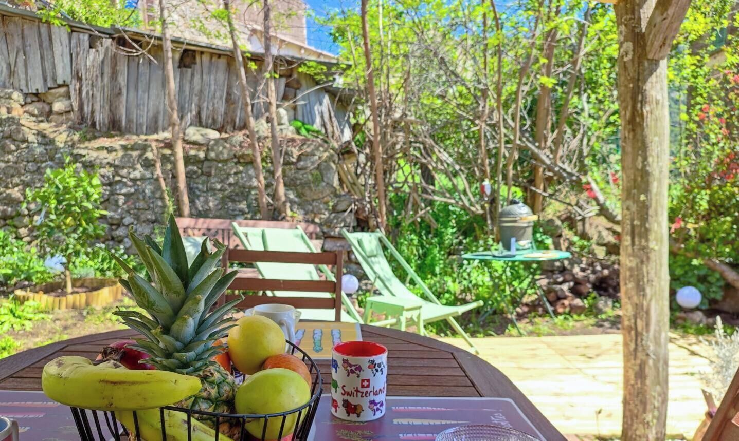 Une table avec des fruits et une tasse, des sièges en plein air, et un mur de pierre en arrière-plan.