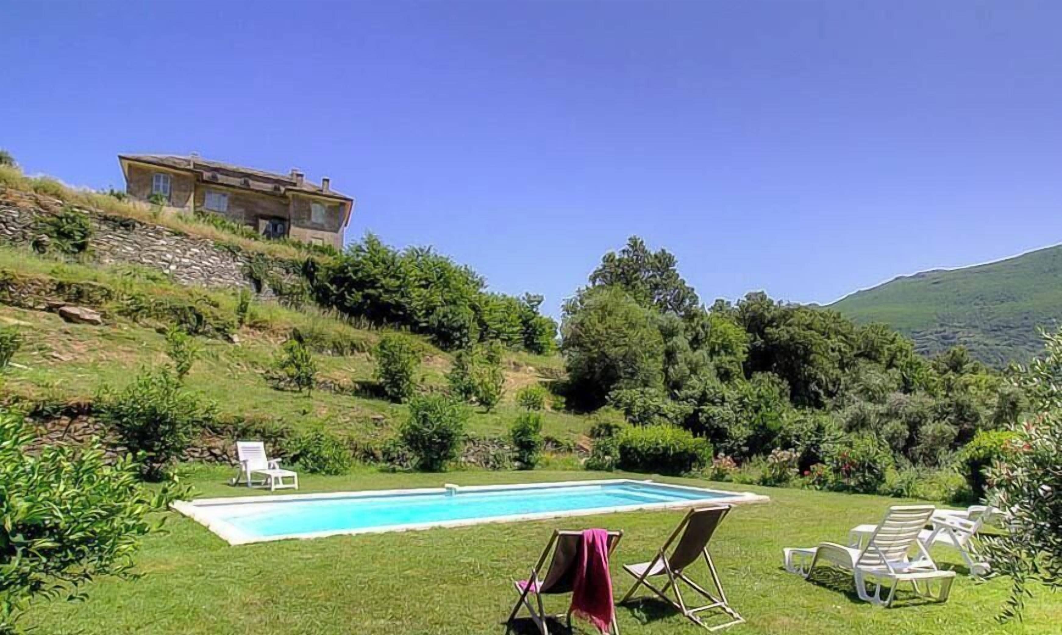 A swimming pool with lounge chairs, a grassy area, and a hillside with a house.