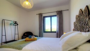 Habitación de hotel con cama, ventana con cortinas, cabecero, estante montado en la pared y lámpara colgante.