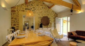 Un coin repas avec une table en bois dressée pour six personnes, des murs en pierre, un luminaire suspendu et un canapé au premier plan.