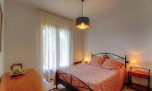 Ein Schlafzimmer mit Bett, einem Tisch mit Pflanze, einem Fenster mit Vorhängen und einer Pendelleuchte.