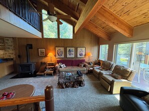 A spacious living room with a high ceiling, wooden beams, a fireplace, and large windows offering views of the outdoors. - Largest Townhome in Leland Meadows, Plus a Hot Tub! (Pinecrest)