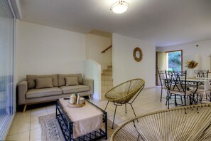 Una sala de estar con sofá, sillas de mimbre, una mesa de comedor con sillas, una escalera y vistas por una ventana.