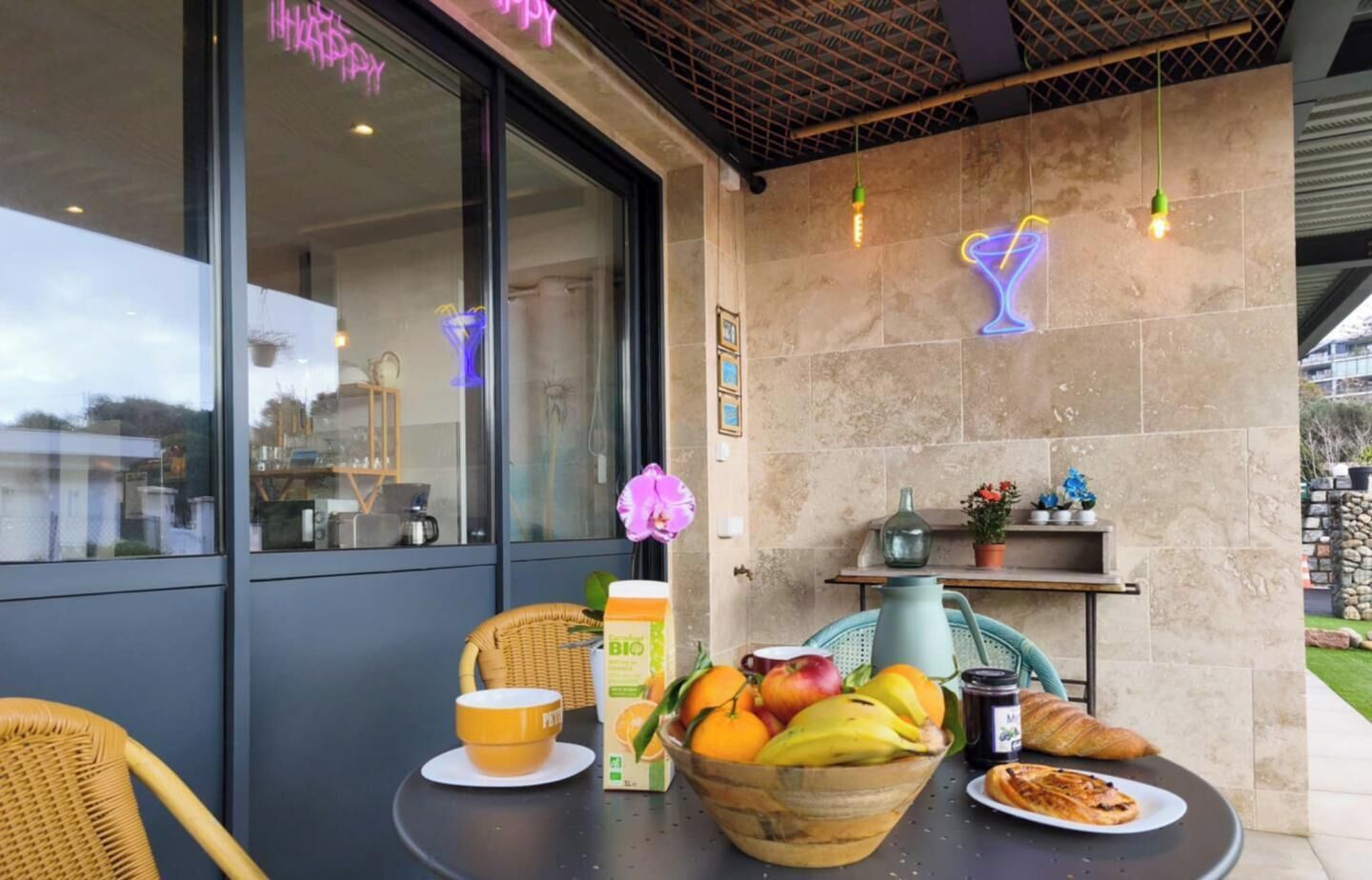 A table with a basket of fruit, a coffee cup, a bottle, and a croissant, set against a modern building facade with large windows and decorative lighting.