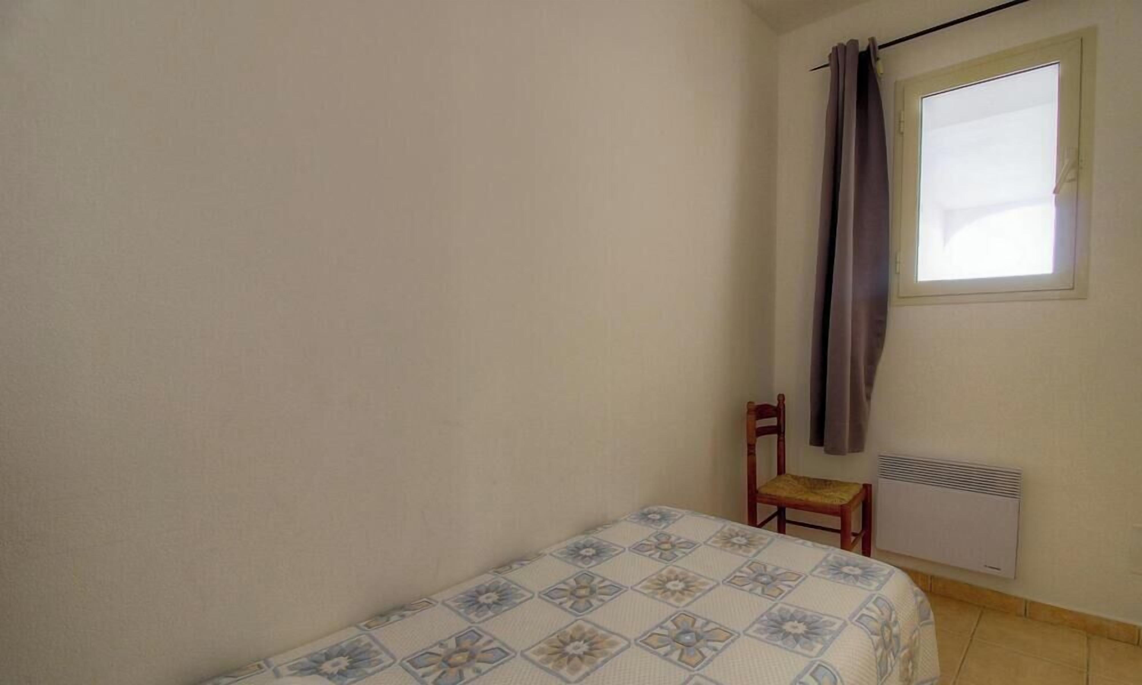 A small, single-bed room with a patterned bedspread, a chair, a window with curtains, and a wall-mounted heater.