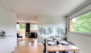 Et moderne spiseområde med et bord dækket til fire personer, et køkken og udsigt til grøn natur.
