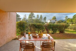 Eine Terrasse im Freien mit einem Tisch für zwei Personen, umgeben von Grünpflanzen und Blick auf Bäume und ein Gebäude.