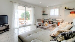 Een woonkamer met een flatscreen televisie, een bank, een eettafel met stoelen en een schuifpui naar een buitenruimte.
