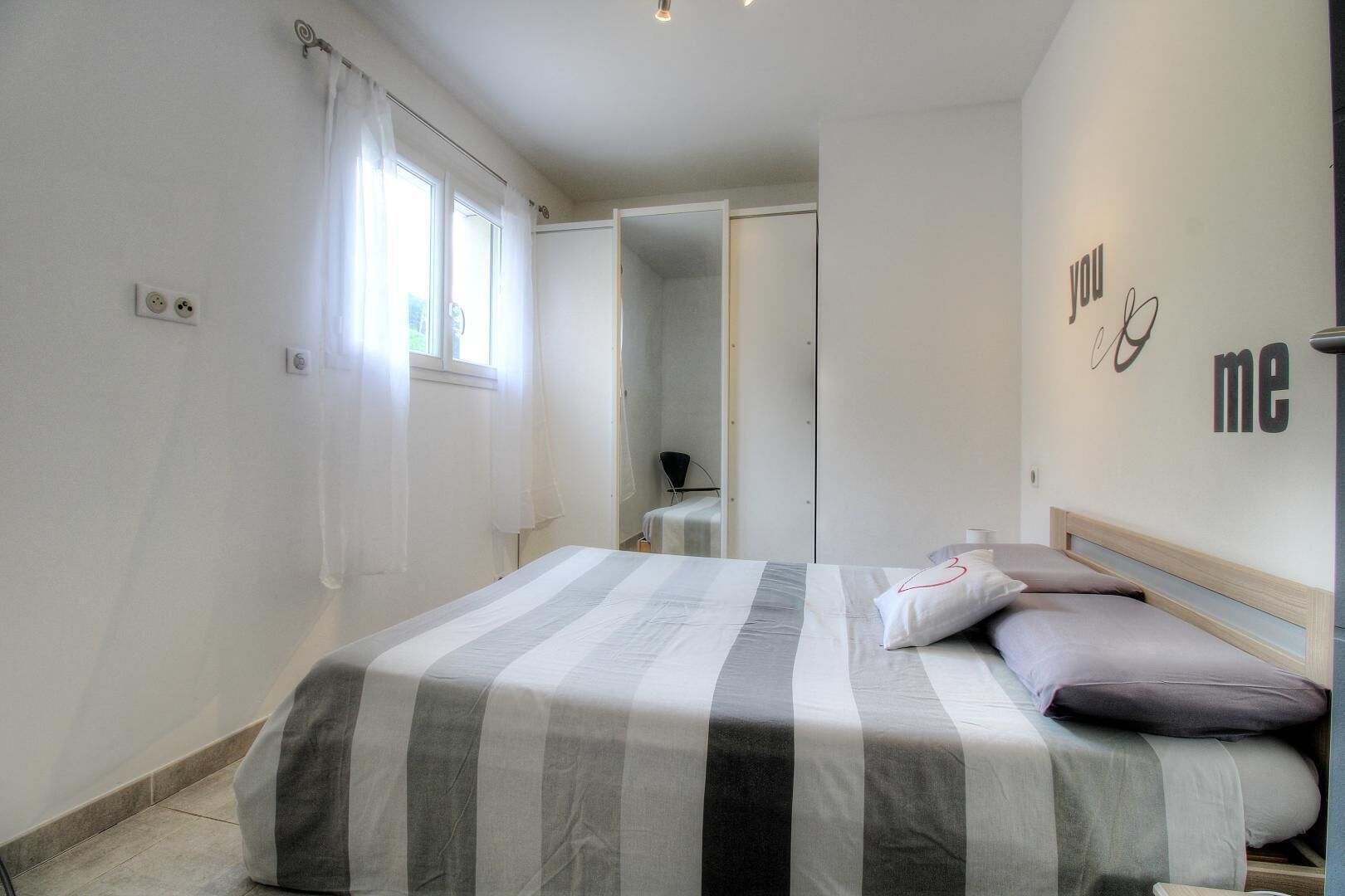 A bedroom with a bed, a bedside table, a mirror, and a wall with 'you me' text.