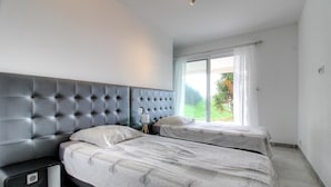 Een moderne slaapkamer met een gestoffeerd hoofdbord, twee bedden, een nachtkastje, een lamp en een groot raam met uitzicht op groen.