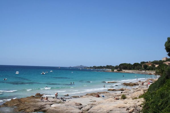 Ein Strand mit felsigem Gelände, klarem blauen Wasser und Gebäuden in der Ferne.