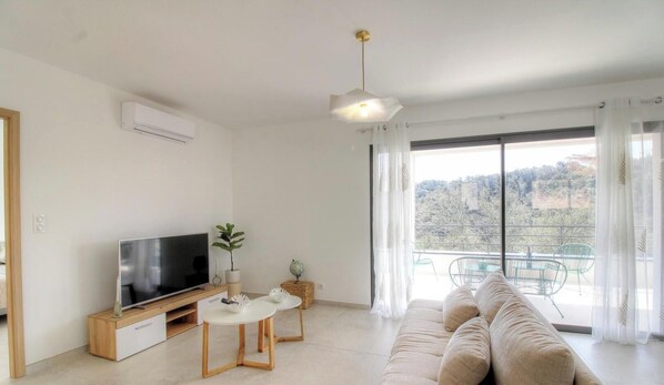 A modern living room with a flat-screen TV, a sofa, a dining table, and a balcony with a view. - Casa Valinina 1 - BASTELICACCIA (BASTELICACCIA)