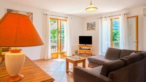 Een woonkamer met een bank, een televisie, een lamp en een tafel. Gordijnen en uitzicht op groen buiten.