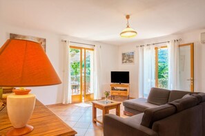 Ein Wohnzimmer mit einer Couch, einem Fernseher, einer Lampe und einem Tisch. Vorhanden sind Vorhänge und ein Blick ins Grüne.