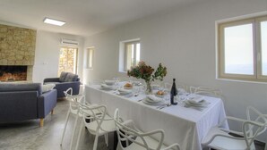 Un coin repas avec une longue table dressée pour six personnes, des chaises blanches, une cheminée en pierre et un canapé gris.